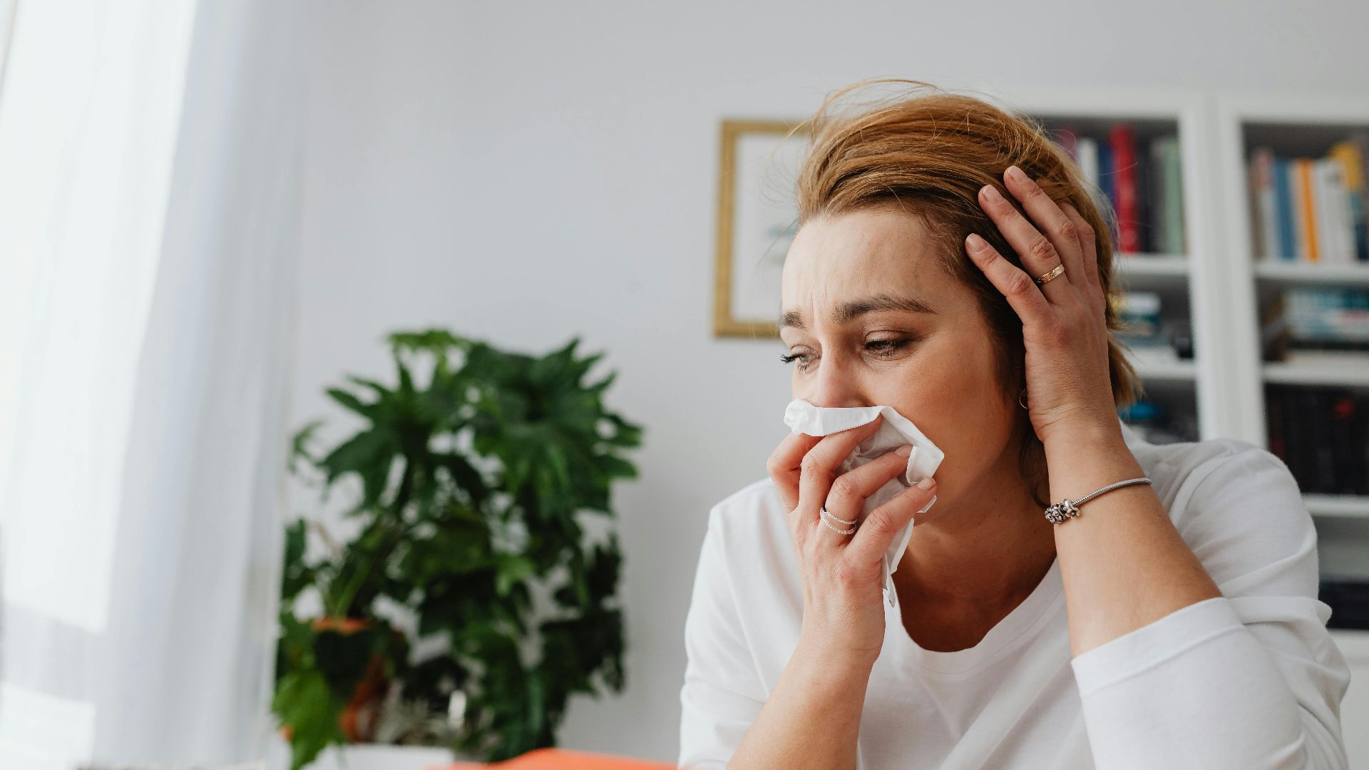 An adult woman feeling unwell, blowing her nose indoors.