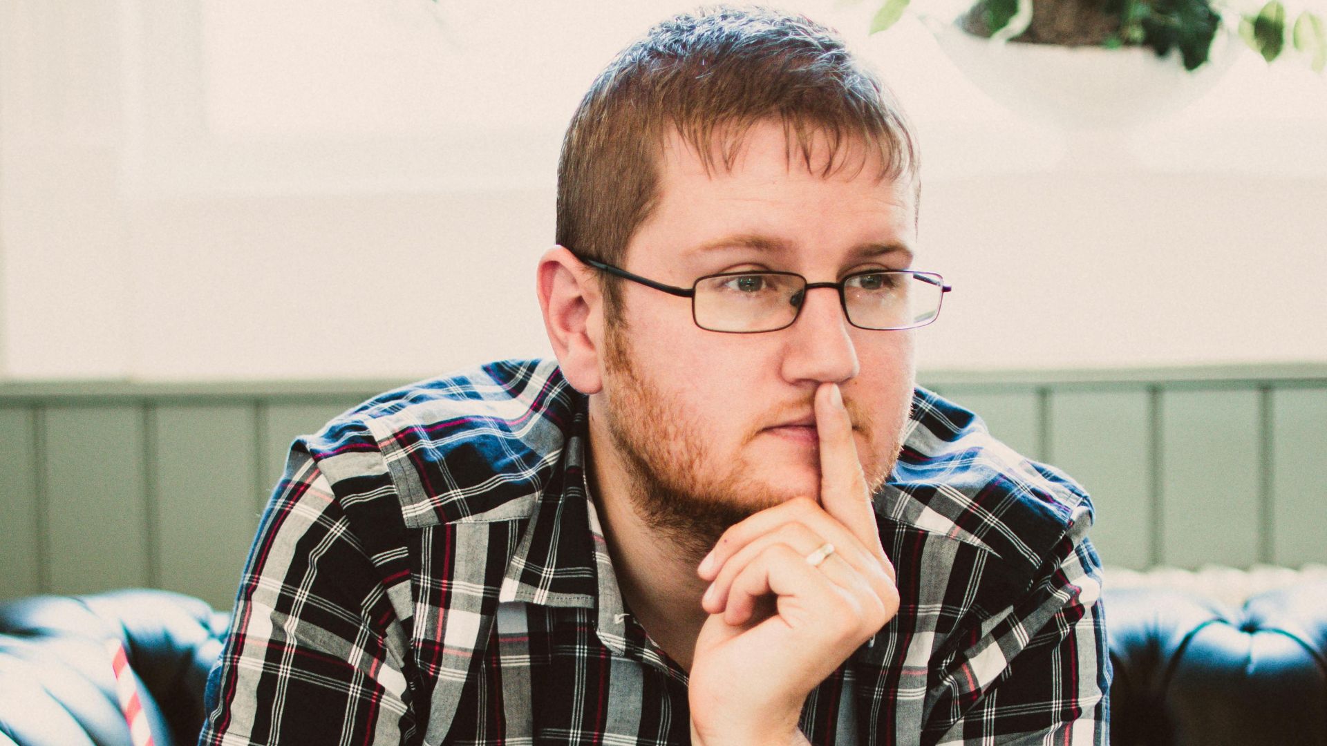 Young man wearing glasses thinking while writing in a cafe with a drink beside him.