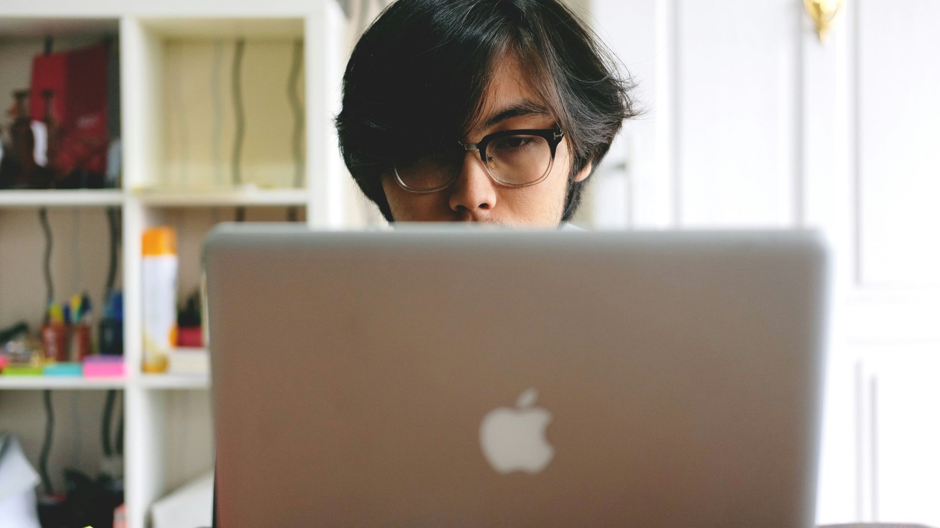 man wearing eyeglasses while using silver Macbook near wooden cubby shelf inside room