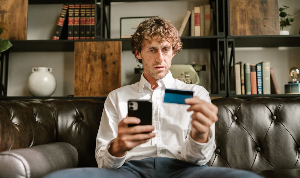 A man sitting on a leather sofa using a smartphone and credit card for online shopping.
