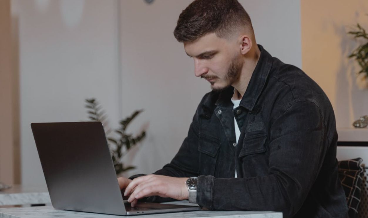 Young man in a denim jacket working on a laptop indoors, showcasing modern remote work lifestyle.
