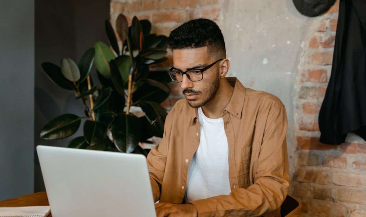 A man in glasses working on a laptop in a cozy cafe with brick walls and plants.