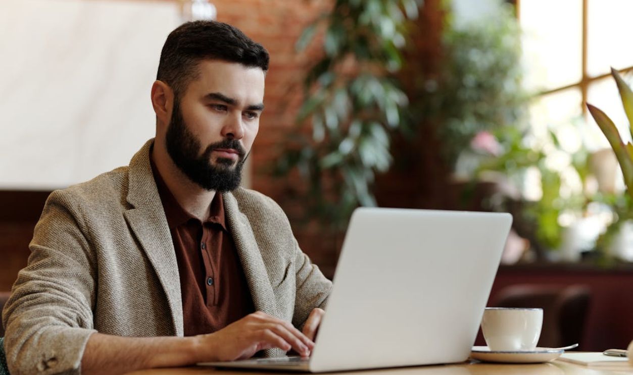 Businessman working on laptop in a cafe setting, surrounded by plants, sipping coffee.