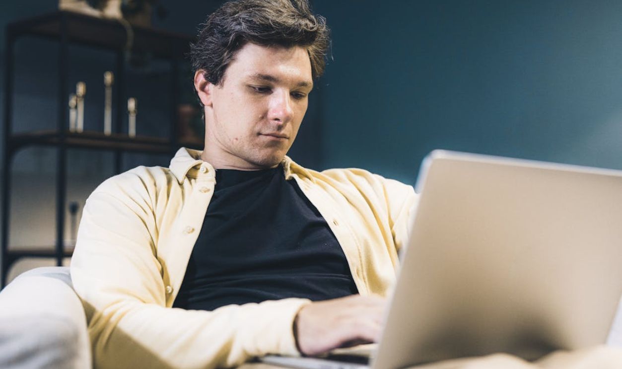 Focused young man sitting comfortably using a laptop at home, engaging in remote work.