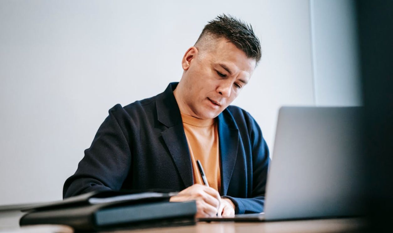 Man engaged in work, writing notes at a desk with a laptop.