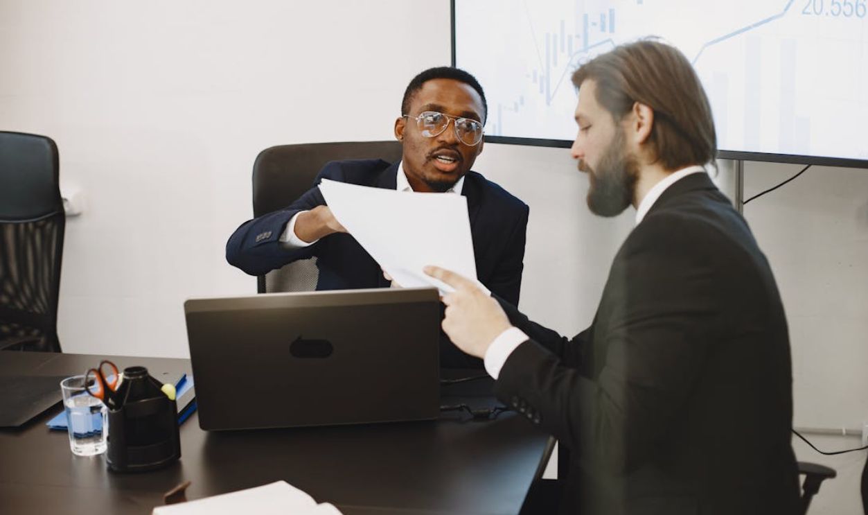 Two businessmen in suits discussing documents at an office meeting with a laptop and charts.