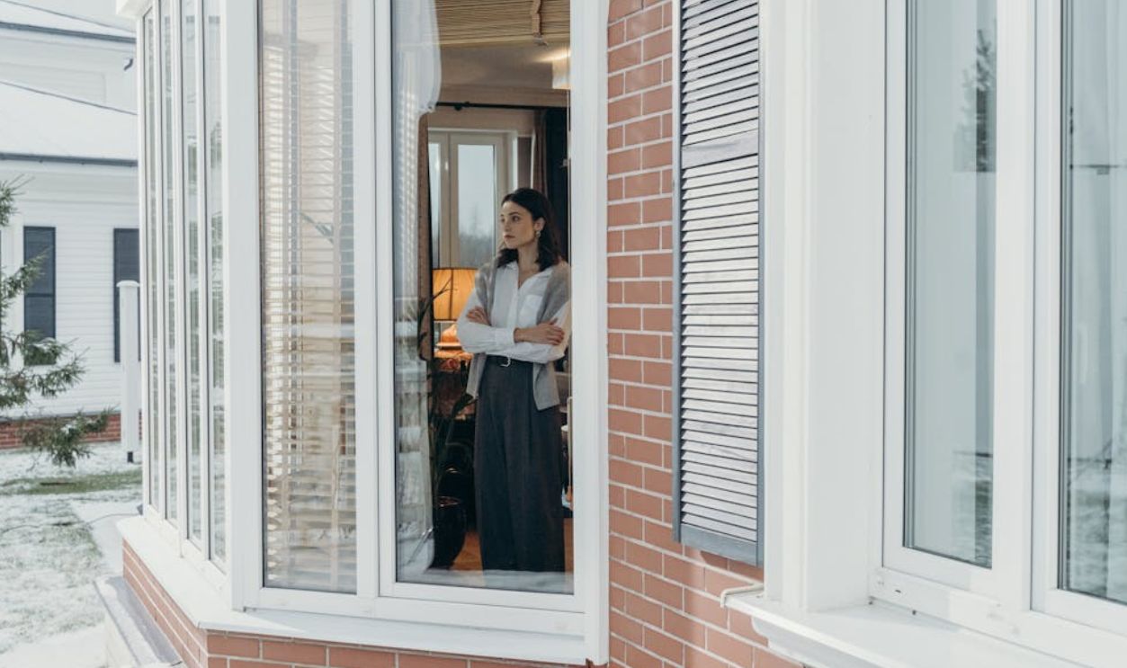 A woman stands indoors, arms crossed, looking out large windows on a snowy day, reflecting indoors and outdoors contrast.