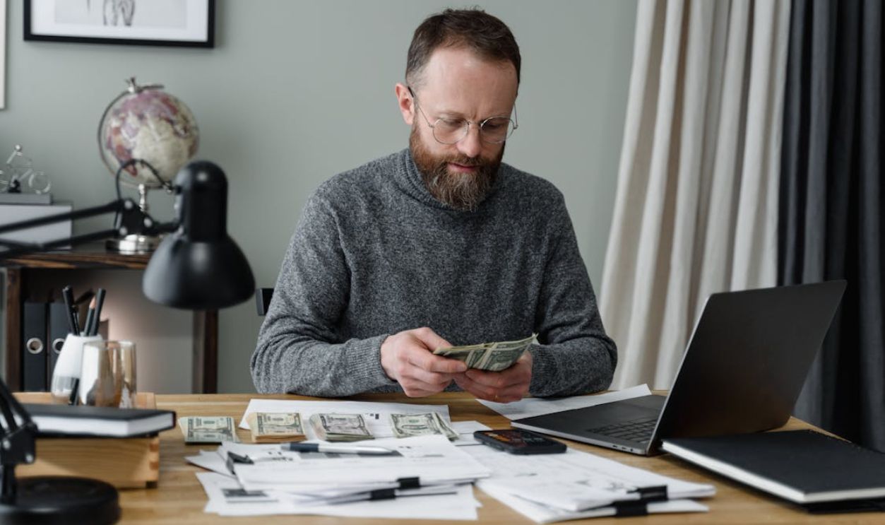 Businessman counting cash at a desk with documents and a laptop in an office.