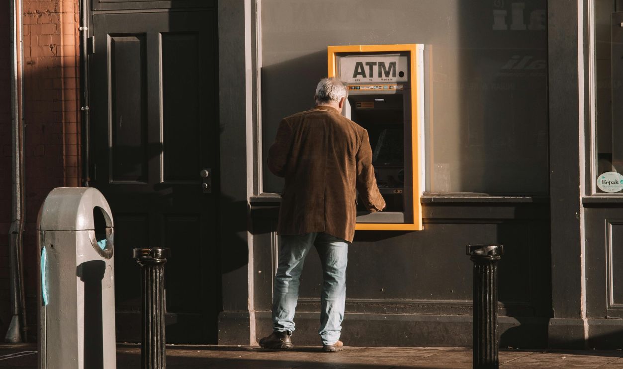 A man using an ATM on a city street during daytime, captured in a vertical shot.