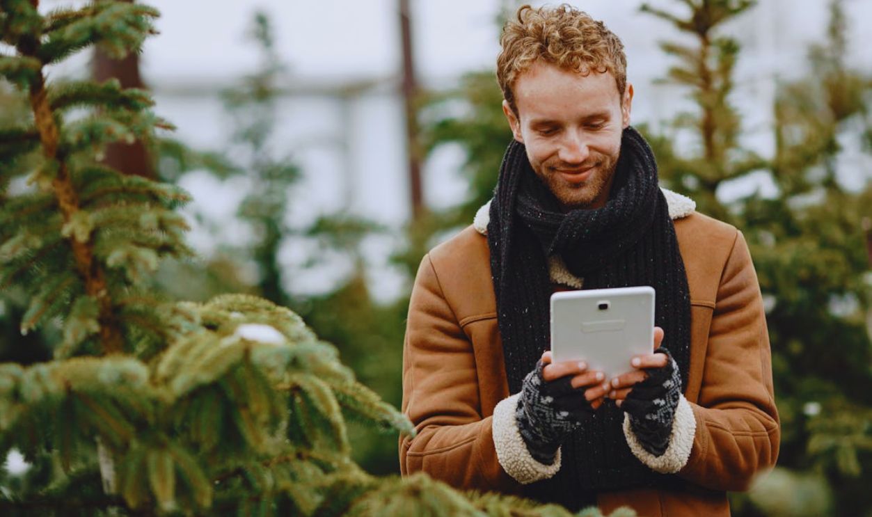 Man in a brown jacket using a tablet in a forest setting during winter, smiling