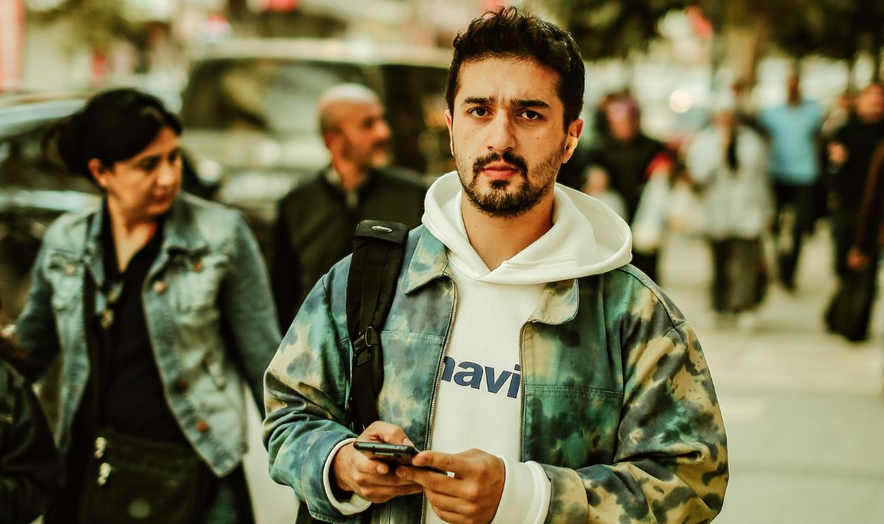 A man in a camouflage jacket walks down a busy urban street, surrounded by pedestrians
