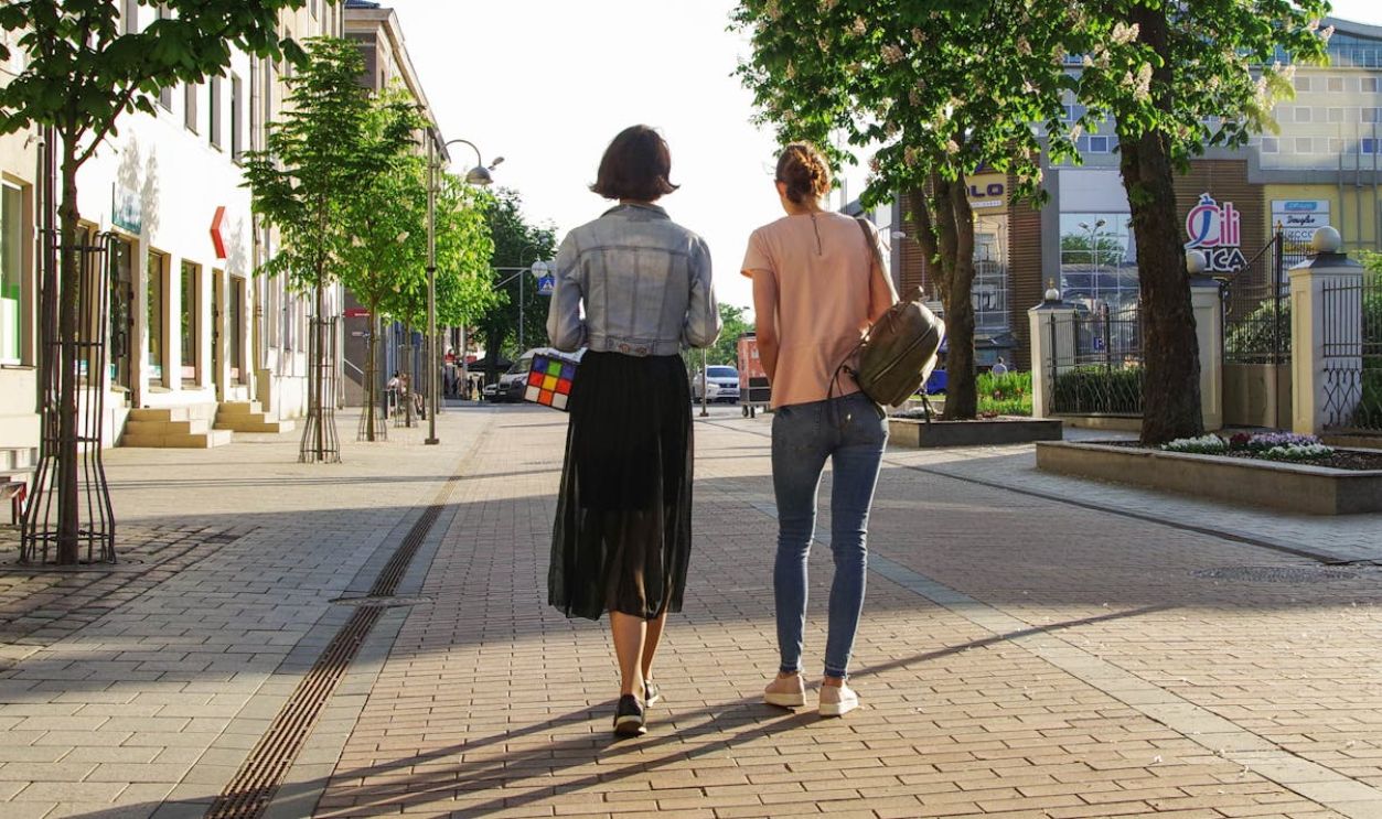 Two women enjoy a sunny walk down a lively urban street lined with buildings and trees