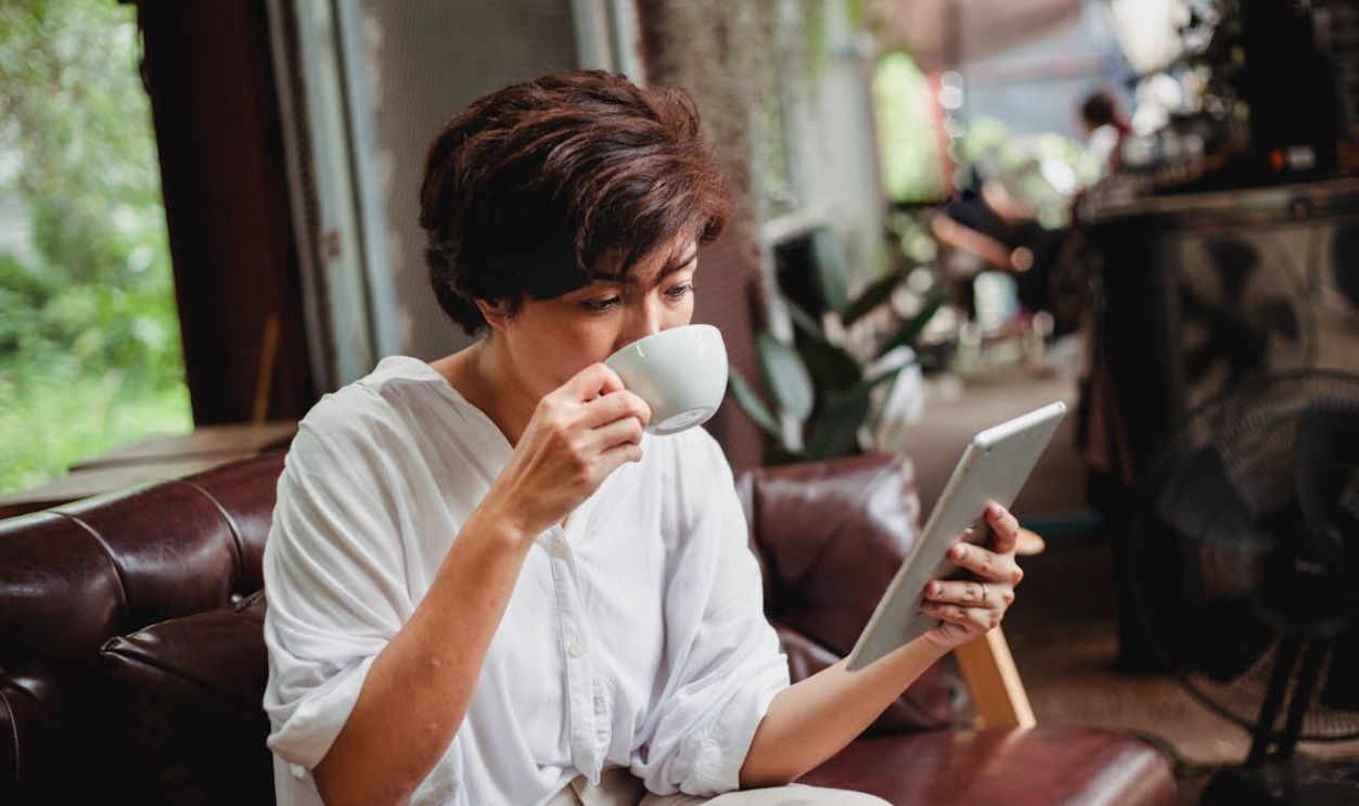A confident Asian woman enjoys coffee while browsing a tablet indoors, conveying focus and relaxation
