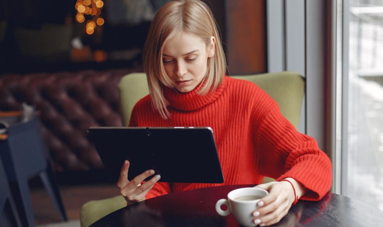 Woman in a red sweater sitting and using a tablet with a cup of coffee in a cozy cafe