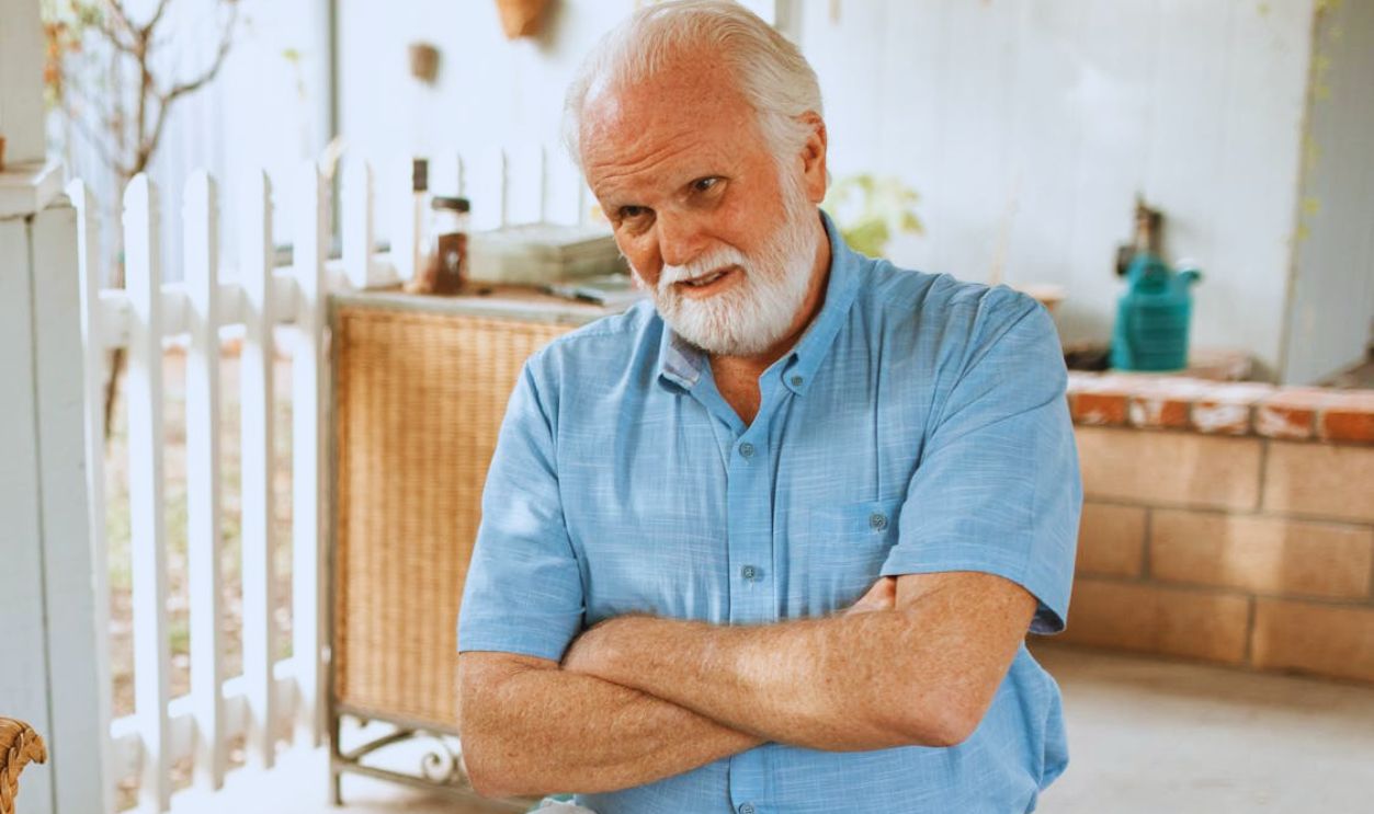 Senior man with white hair and beard sitting on a porch, arms crossed, in a relaxed setting