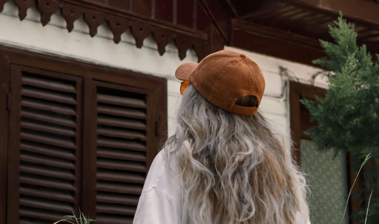 Back view of a woman with gray hair and cap, observing a rustic wooden house facade.