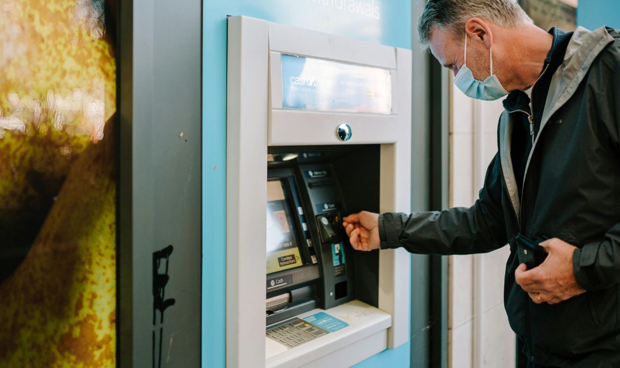 A man wearing a mask using an ATM machine outdoors for cash withdrawal