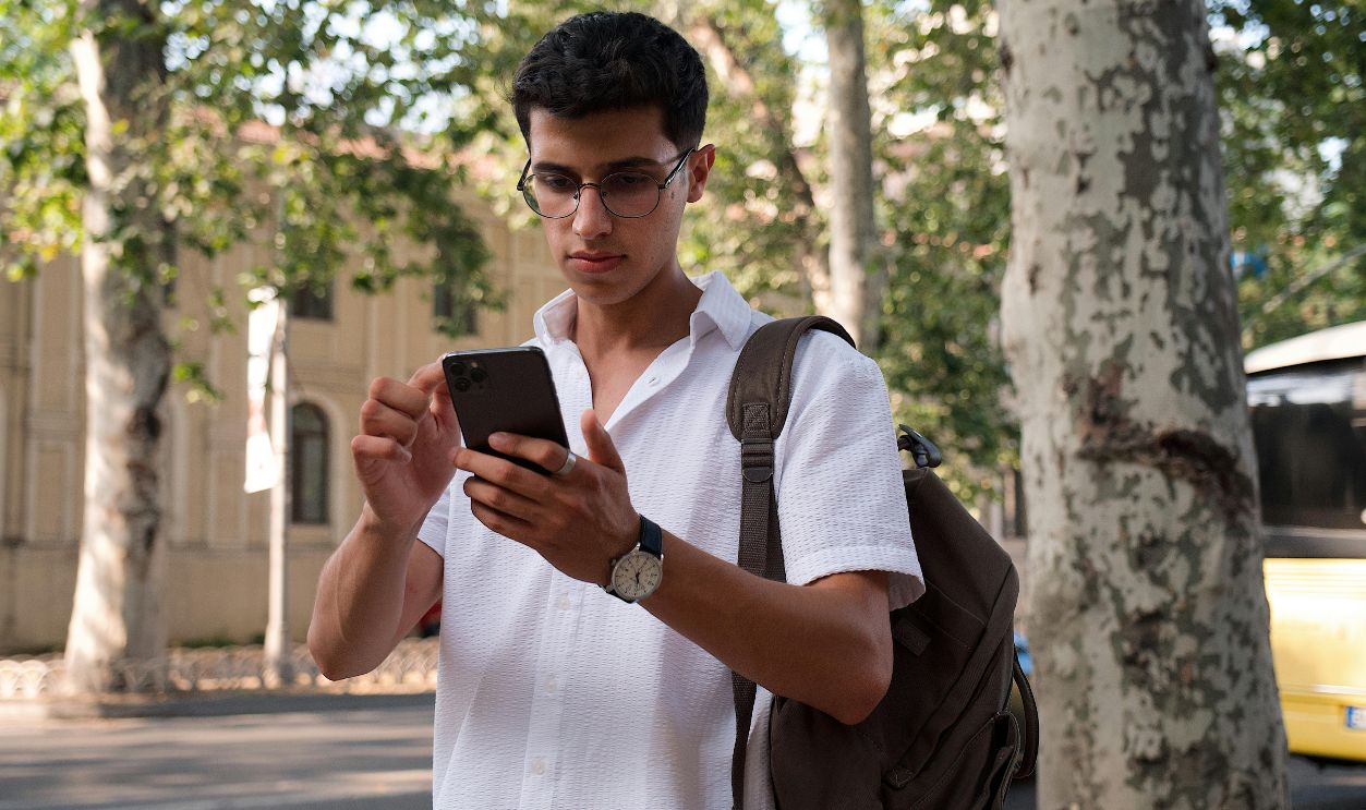 Young man using smartphone on a city street, wearing eyeglasses and a backpack