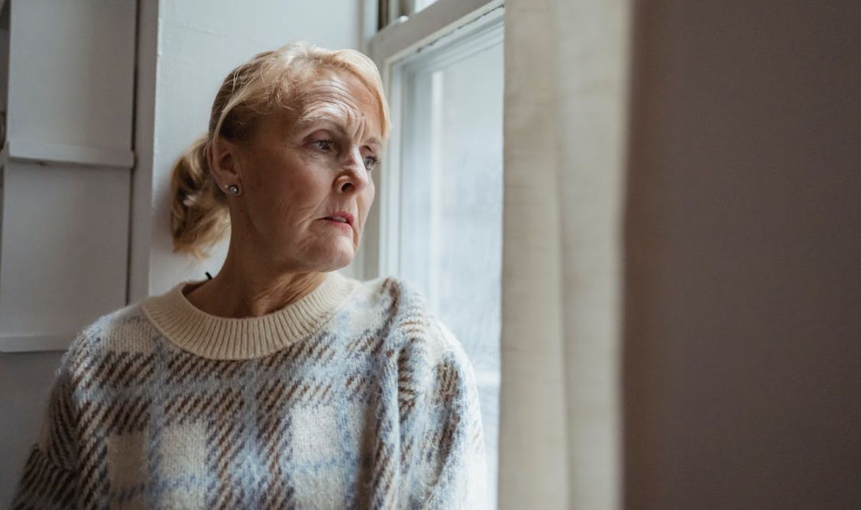 A senior woman in deep thought gazing out a window indoors.