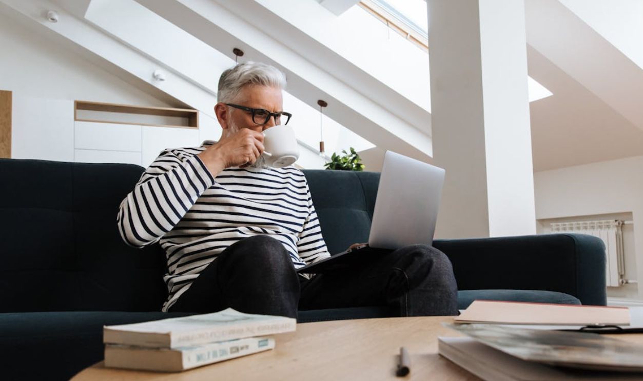 A senior man enjoying coffee while working on a laptop from home in a modern indoor setting.