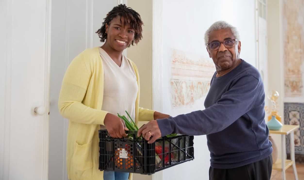 Smiling woman assists a senior man with food delivery indoors, promoting community support and care.