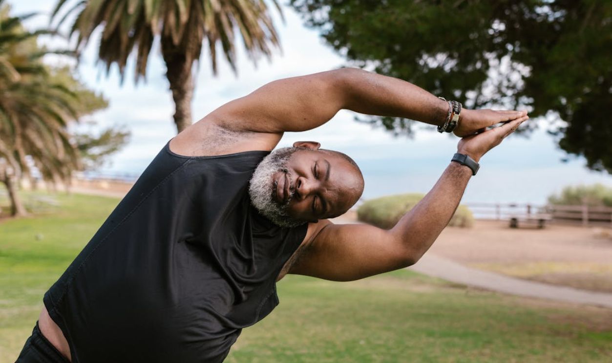 A bearded man in a black tank top stretches in a serene park environment.