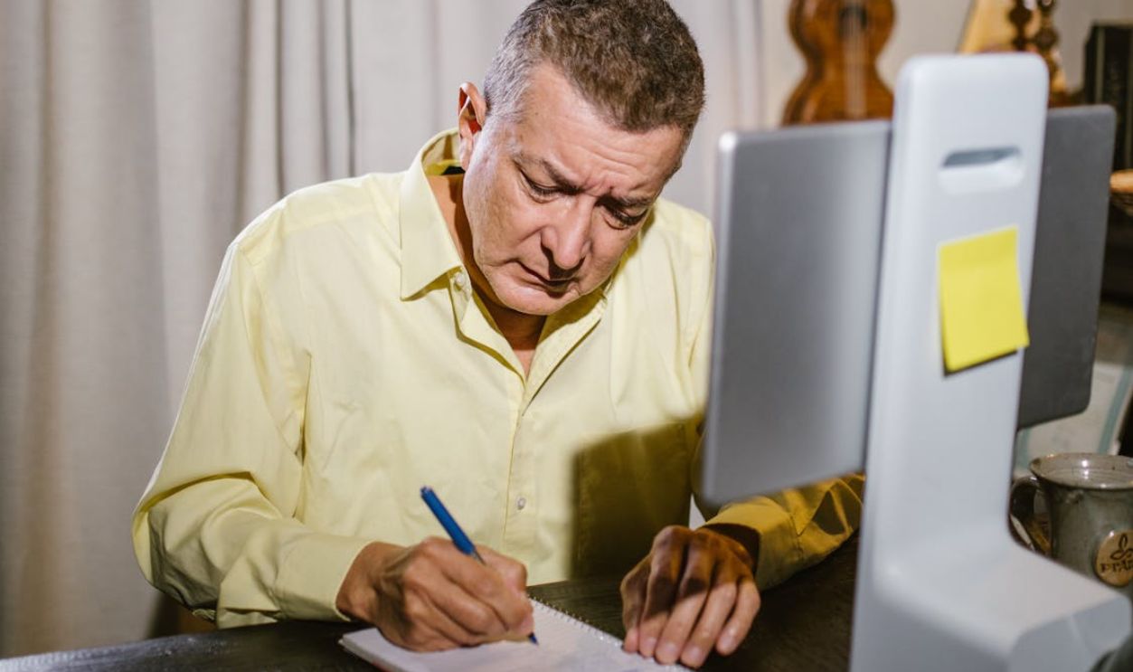 Elderly man focused on writing at home, sitting at a desk with a digital tablet.