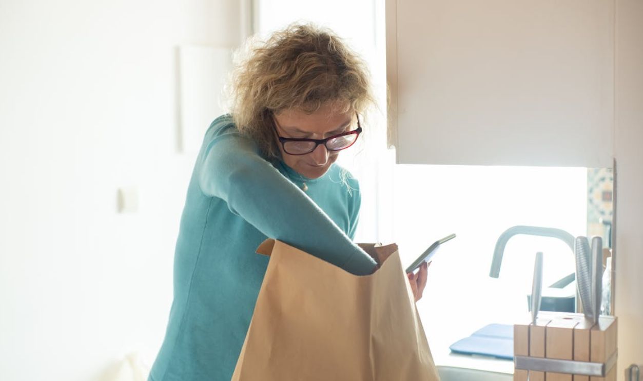 Elderly woman unpacks groceries from paper bag at home, holding smartphone.
