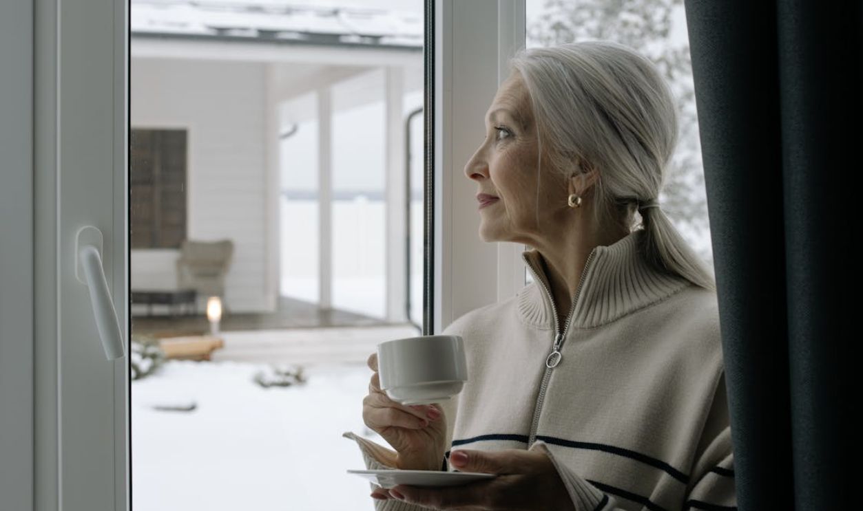 Elegant senior woman with gray hair holds a coffee cup, gazing out a window during winter.