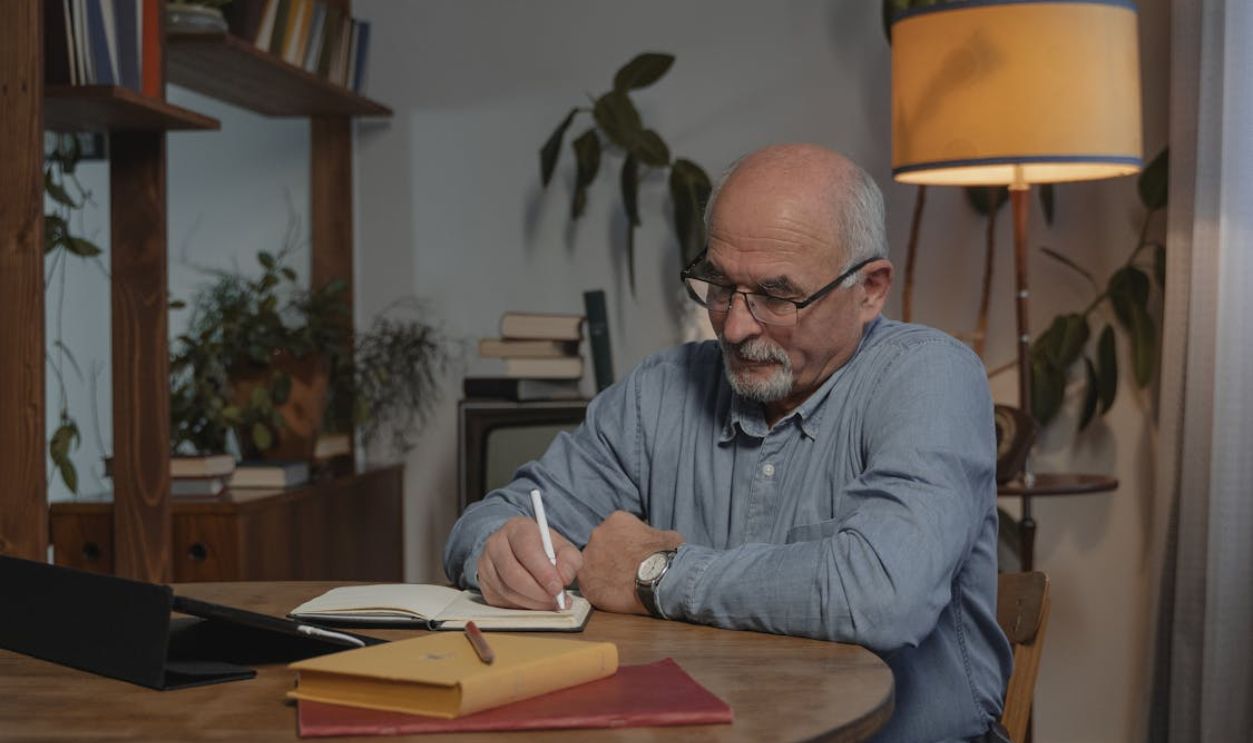 Elderly man pens thoughtful entries in his journal while sitting at a cozy home table.