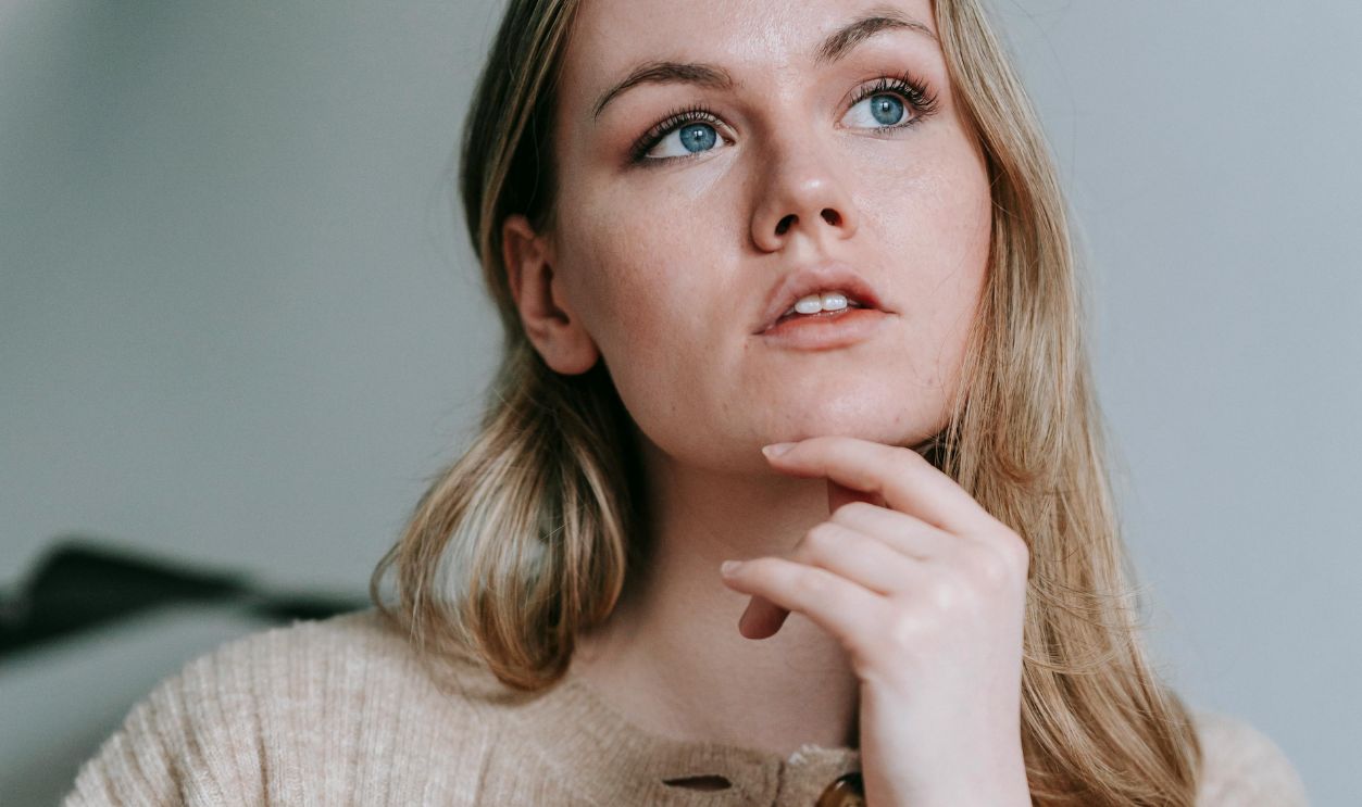 A young woman in a beige sweater sits indoors, thoughtfully looking away while touching her chin