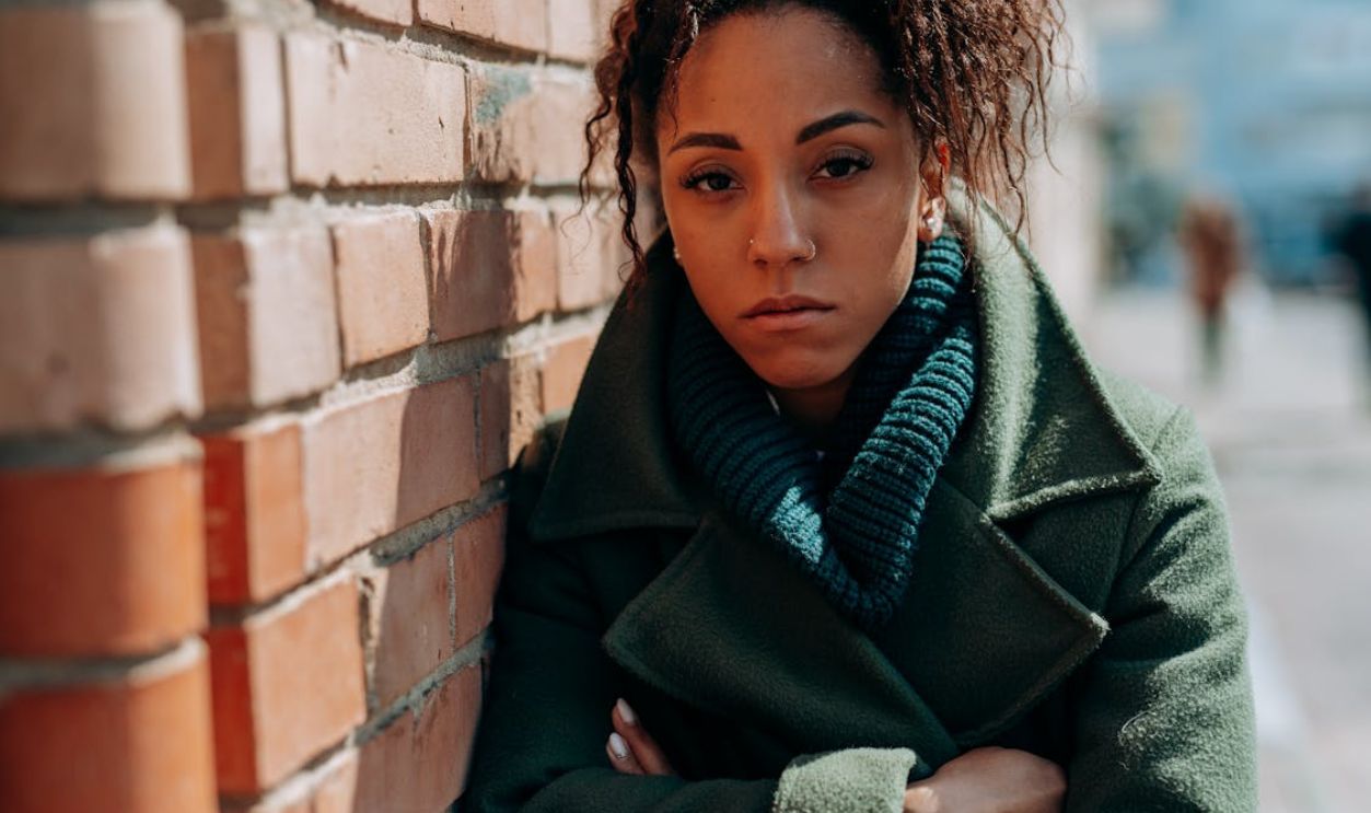 Portrait of a woman in a green coat with curly hair leaning against a brick wall