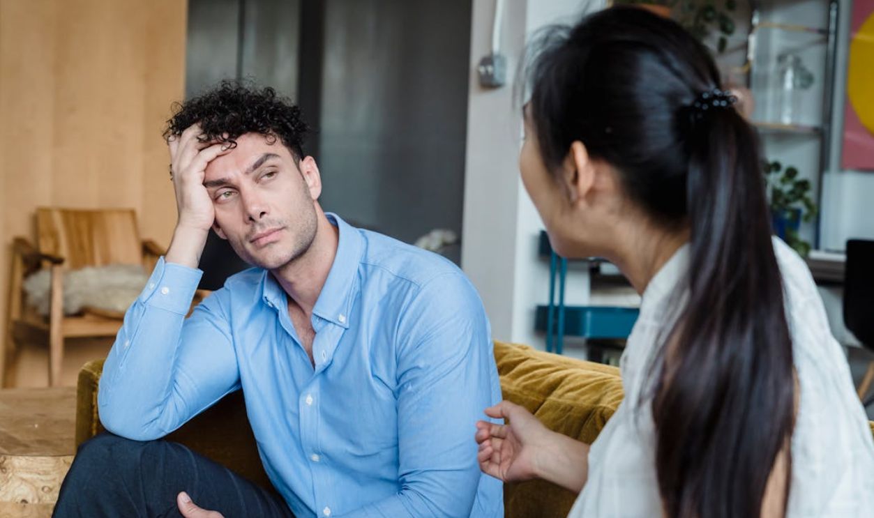 A tired man in a blue shirt listens while a woman talks indoors. Modern living room