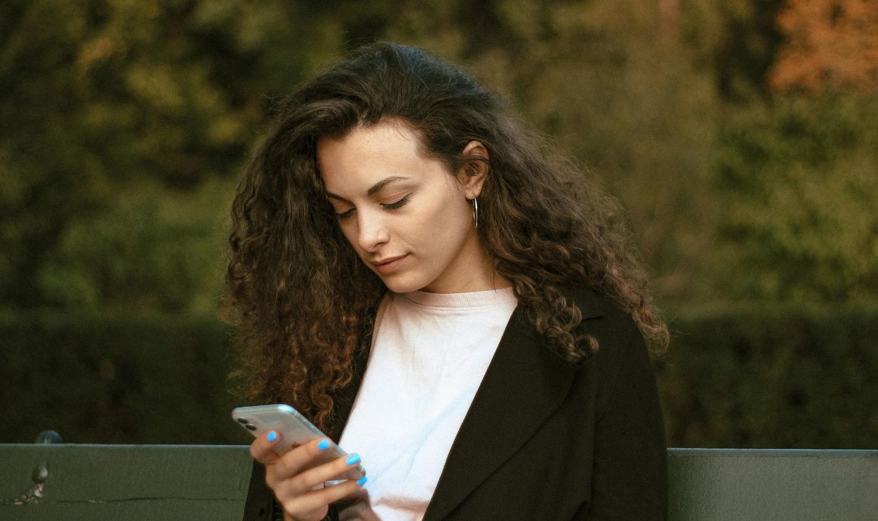 Woman with curly hair checking smartphone while sitting outdoors in Warsaw park