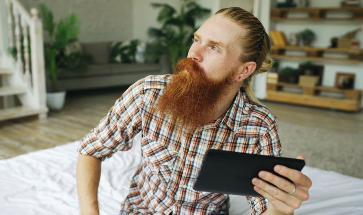 Pensive man with a red beard holding a digital tablet, sitting in a cozy indoor setting.
