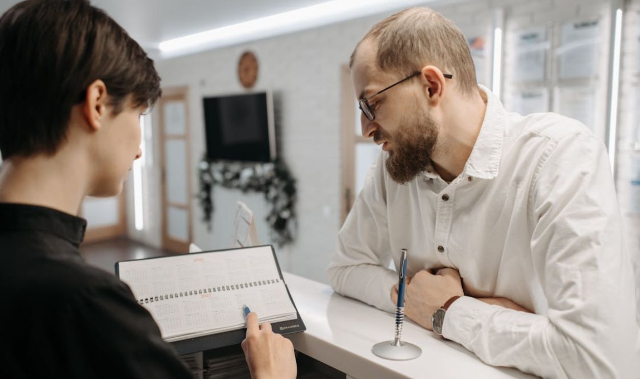 A receptionist and client converse over an appointment book at a clinic reception desk.