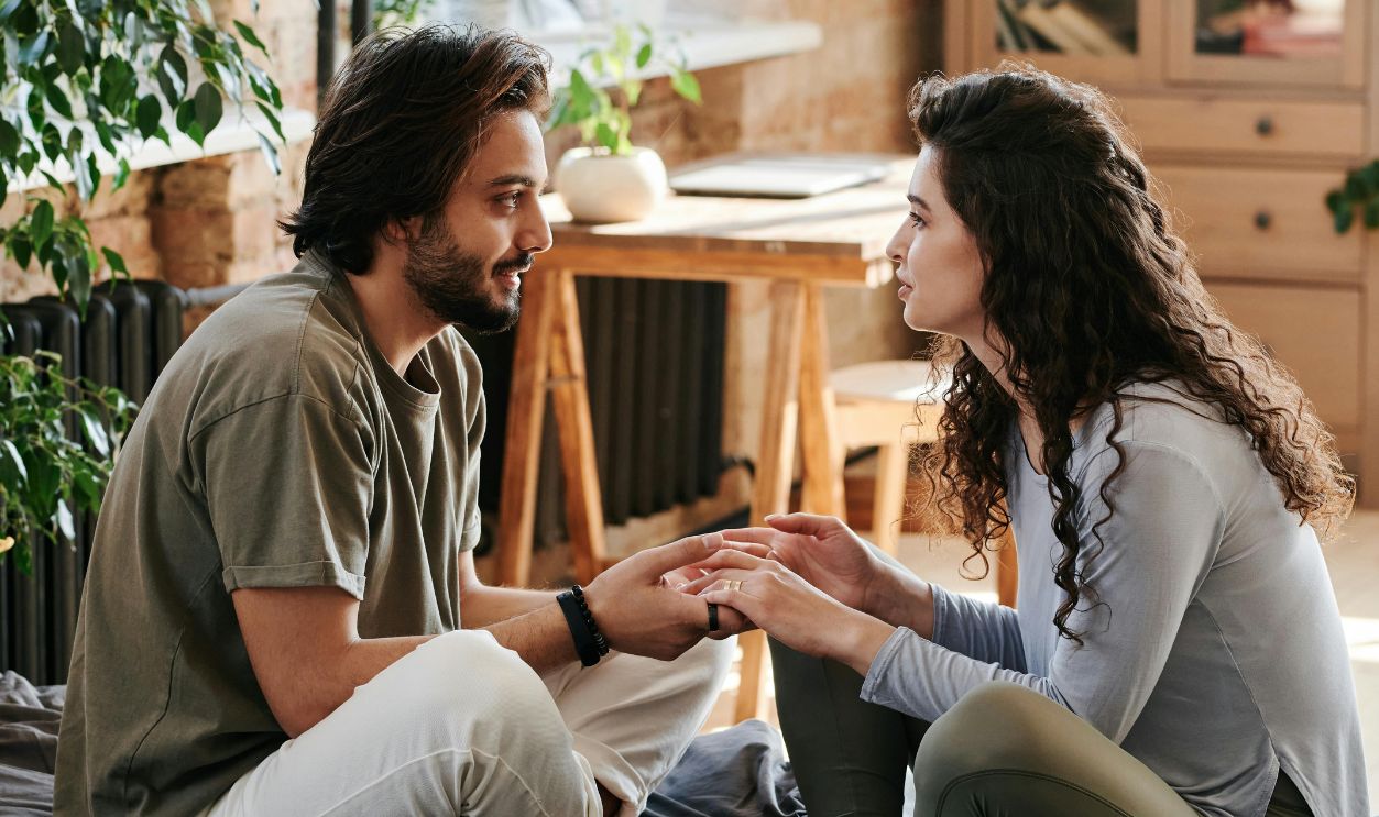 Romantic couple holding hands and talking in a cozy, plant-filled bedroom.