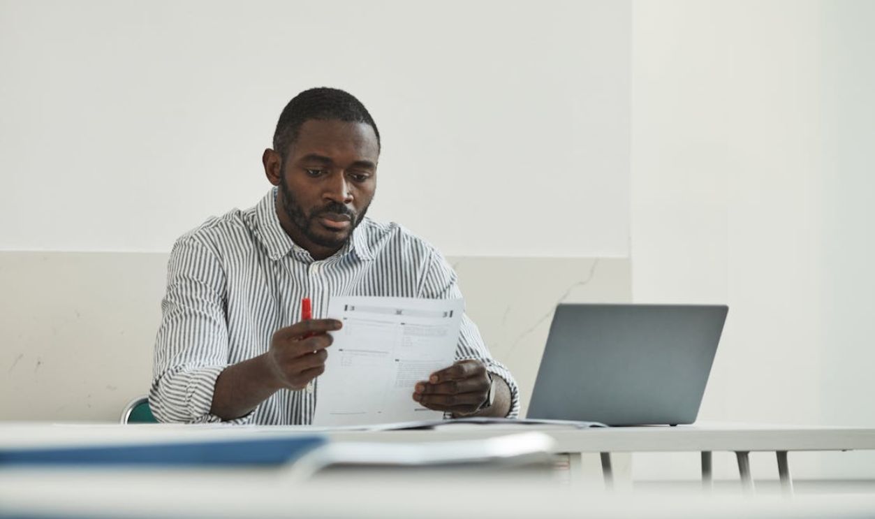 An adult man attentively grading test papers at his workspace with a laptop.