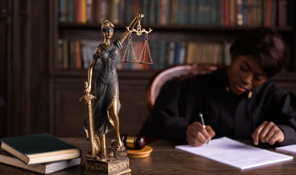 A focused judge writing on documents beside a Lady Justice statue in an office.