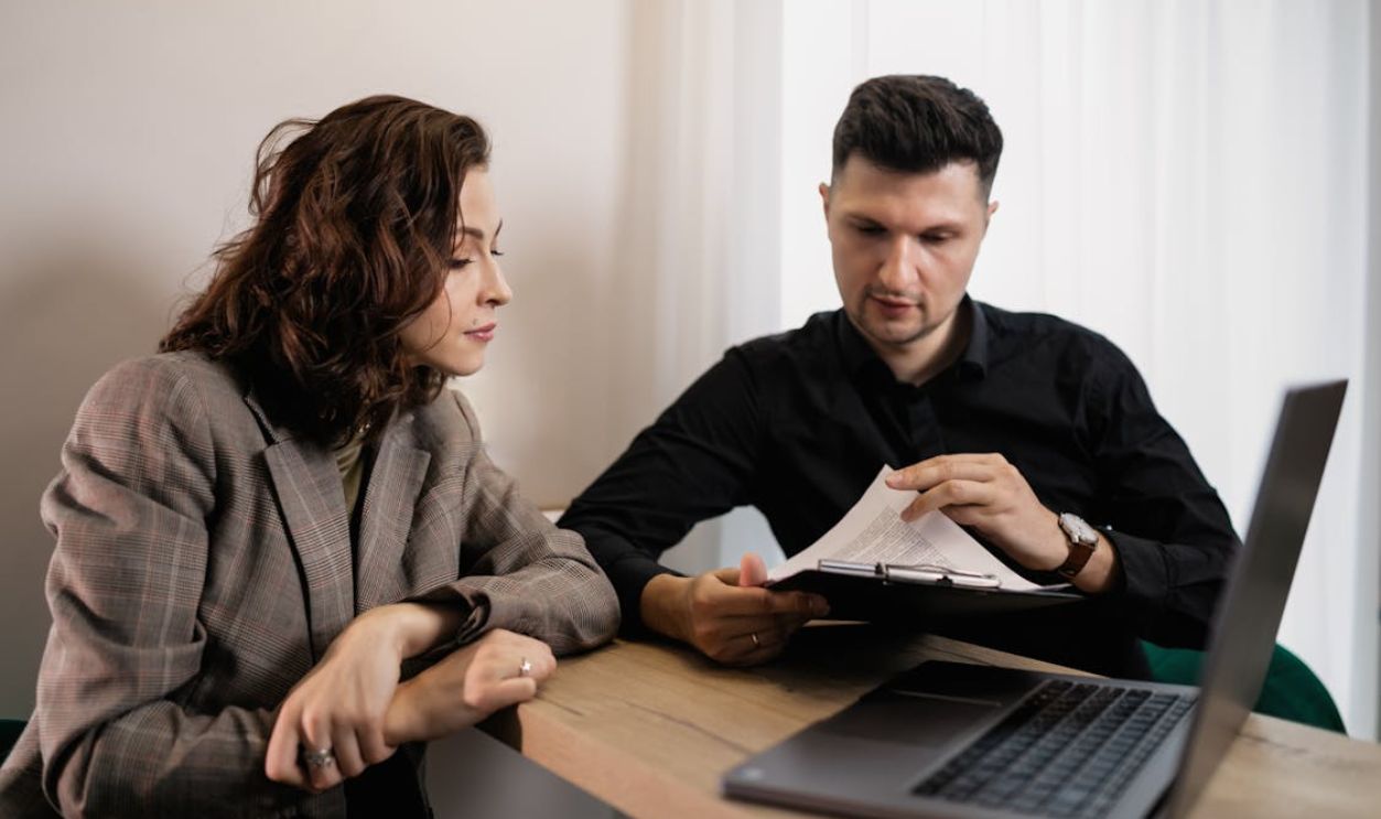 Two professionals discussing documents at a desk with a laptop, focused on collaboration.