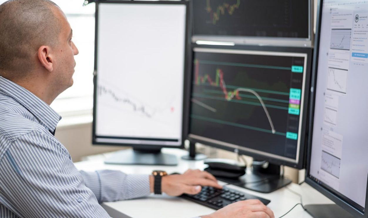 Businessman reviewing financial charts on multiple monitors in an office setting.