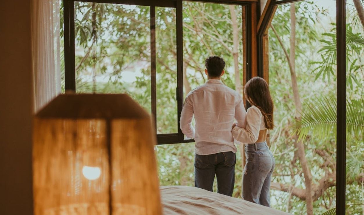 A couple stands by a window in a cozy hotel room with natural lighting and a view of lush greenery.