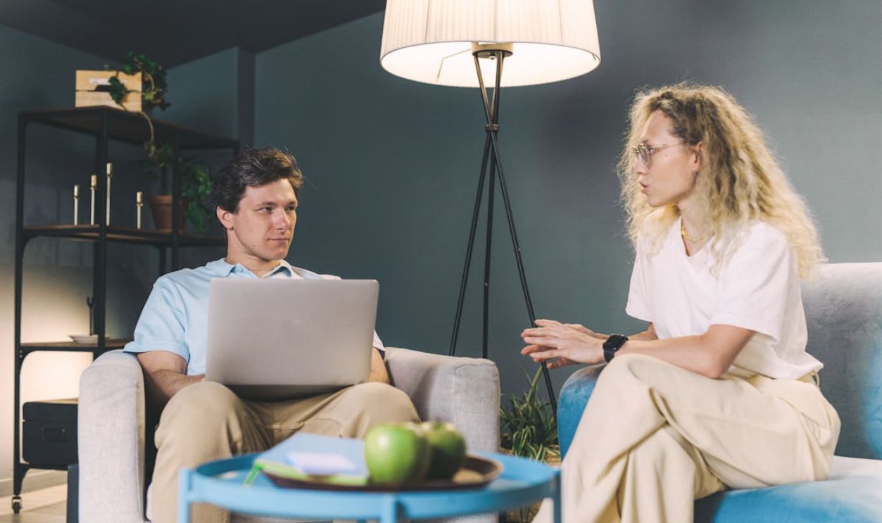 Two adults discuss work while sitting on sofas in a cozy, modern living room setting.