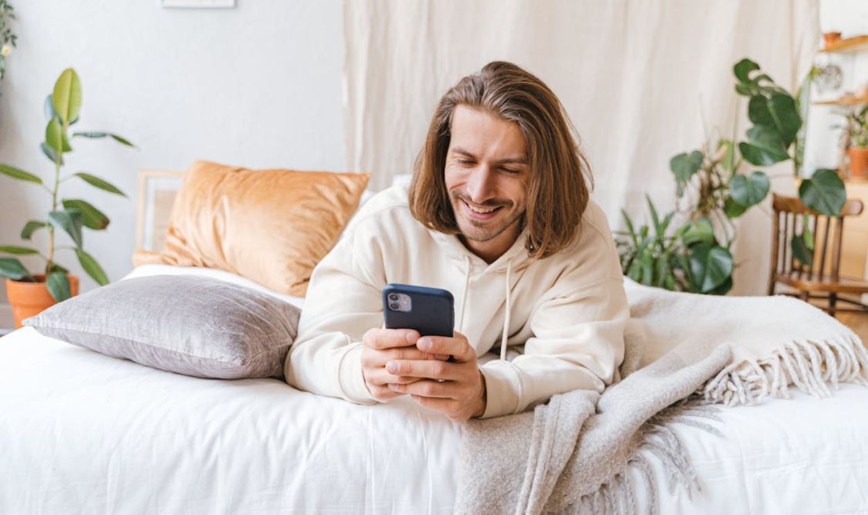 Caucasian man smiling while using smartphone on bed in a bright, cozy bedroom setting.