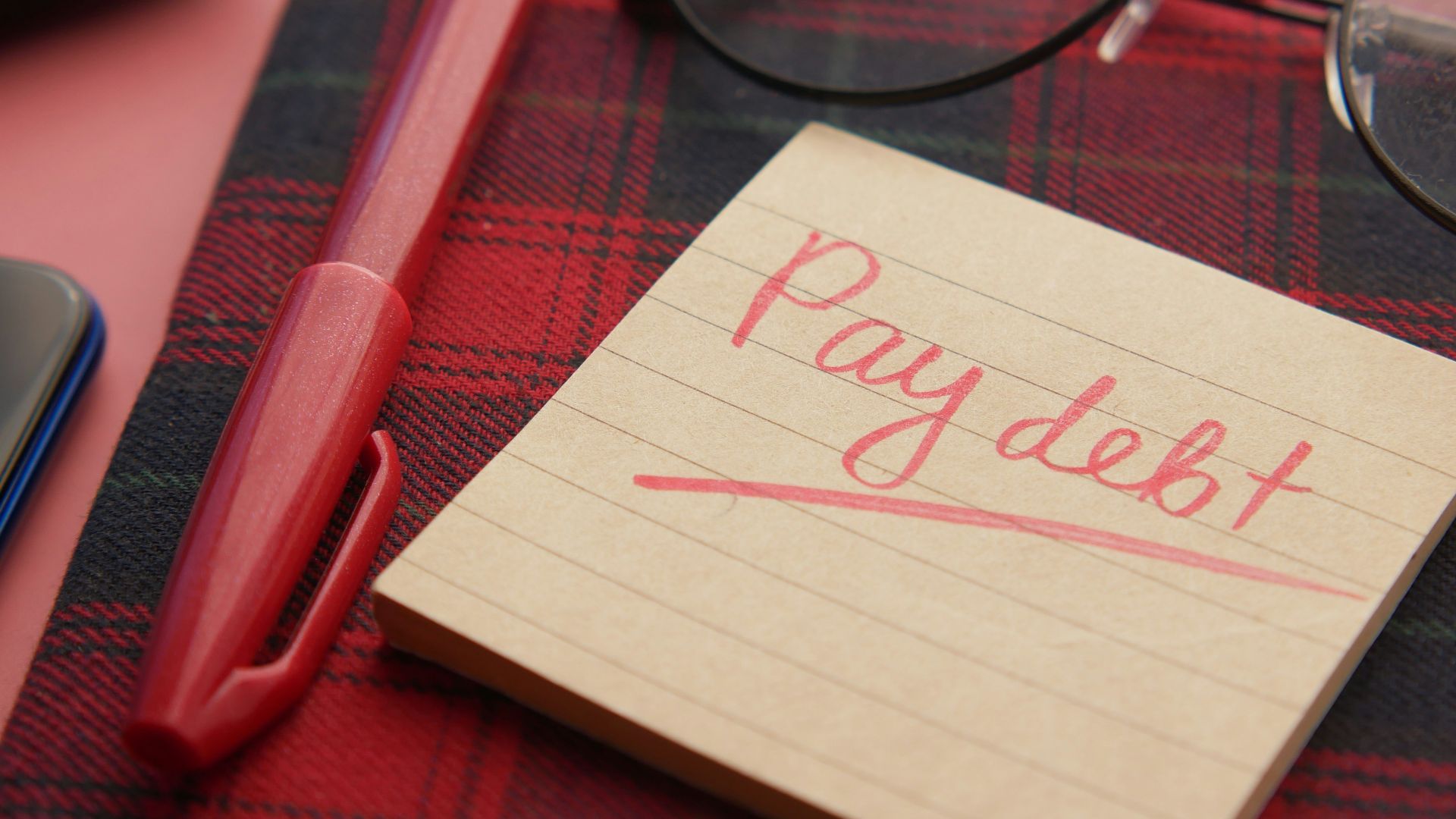 a note that says pay debt next to a pen and glasses