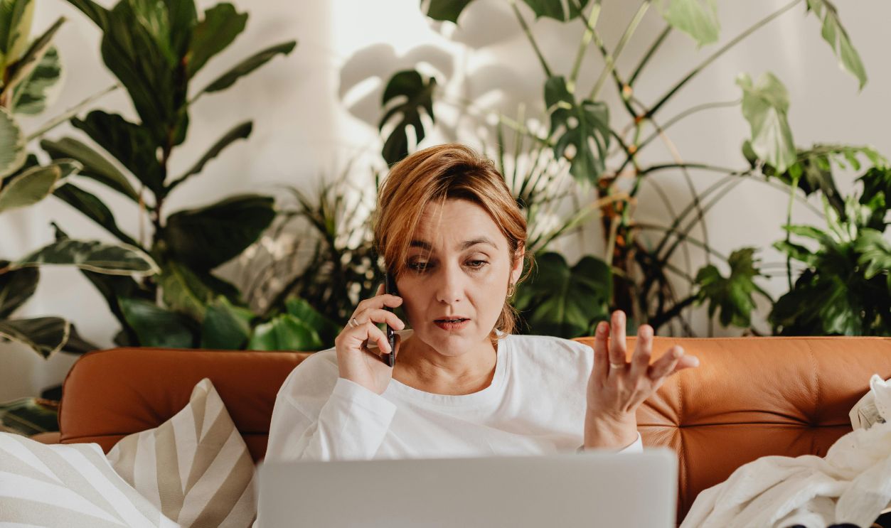 Woman multitasking with laptop and phone on a cozy sofa surrounded by lush indoor plants