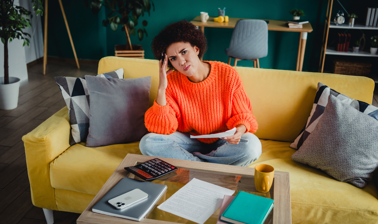 A young woman sits on a yellow sofa at home, looking frustrated while reviewing bills