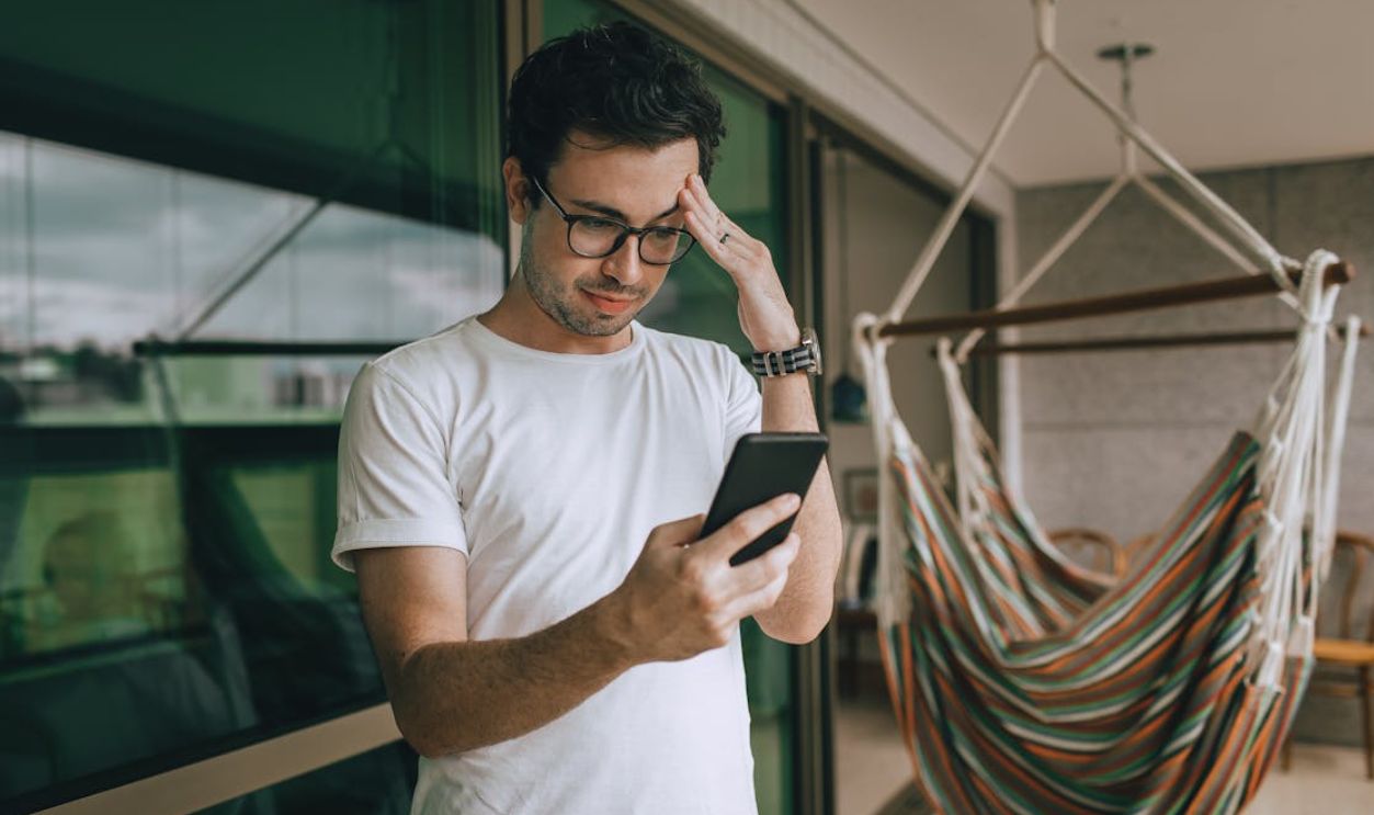 Young man with glasses checking his smartphone by a hammock indoors. Lifestyle and technology theme