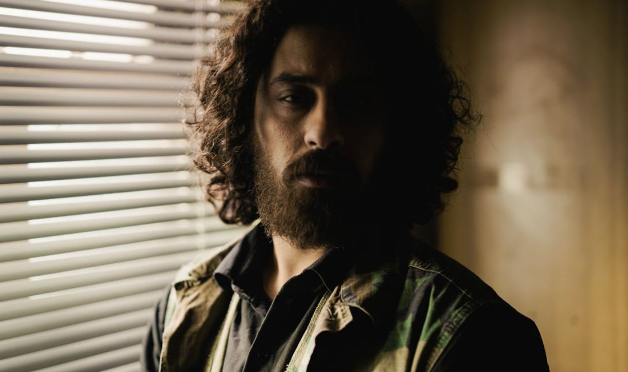 Moody portrait of a man with curly hair and beard standing by window blinds indoors