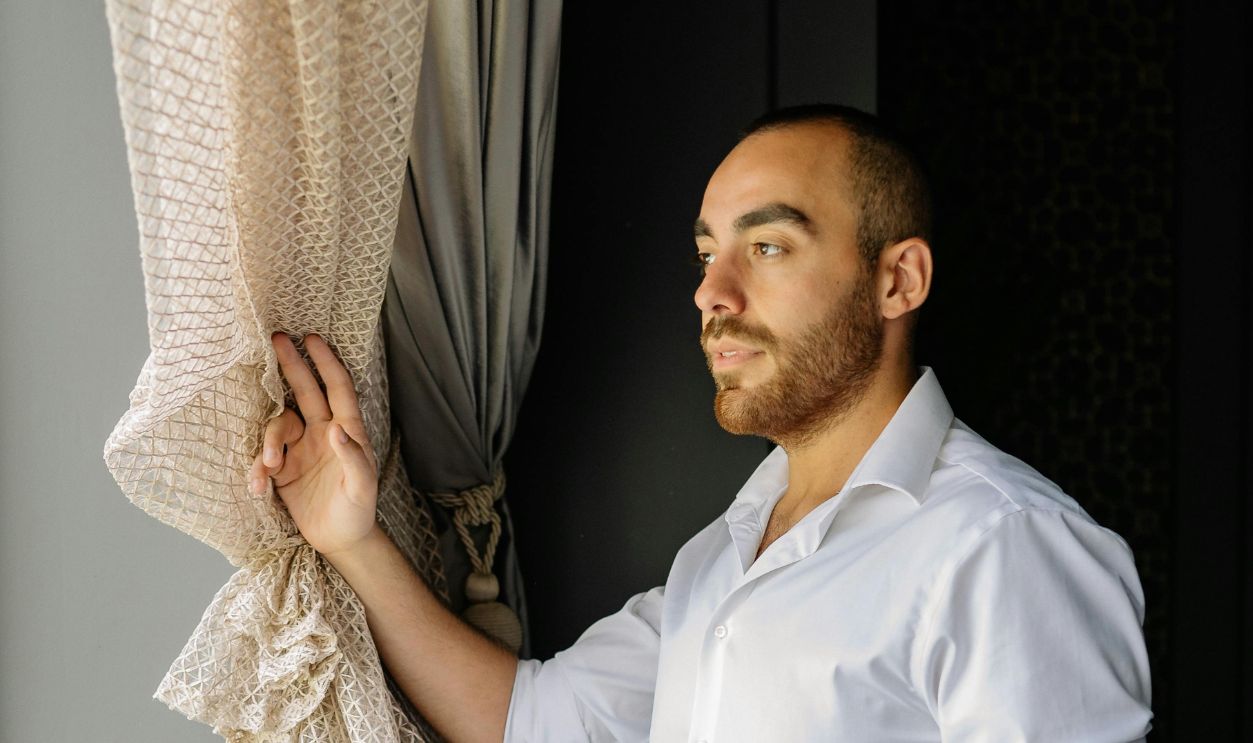 Adult man in a white shirt standing by a window, gently holding a curtain and gazing outside
